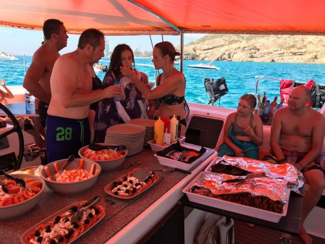 Pasajeros disfrutando de un bufé de barbacoa bajo el toldo del catamarán junto a la costa